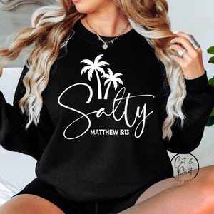 Op de afbeelding: Zwarte sweatshirt met een witte palmboom en het woord "Salty" in sierlijke letters. De tekst "Matthew 5:13" staat eronder. Een comfortabele, casual stijl.