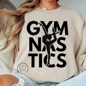 May include: A beige sweatshirt with the text "GYMNASTICS" in black with a silhouette of a gymnast in the middle of the word "NAS". The text "Cut & Print Shop" is in a smaller font at the bottom of the sweatshirt.