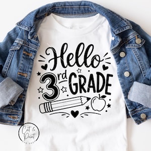 May include: A white t-shirt featuring the black text Hello 3rd Grade, a pencil, an apple, and small heart and star icons, layered underneath a blue denim jacket with metal buttons and cuffed sleeves. The shirt has a round neckline and short sleeves.