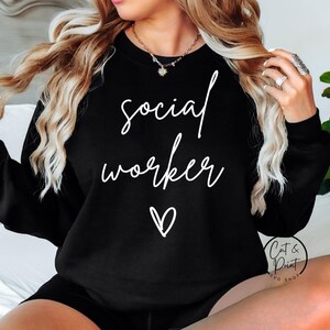 May include: Black sweatshirt with the words "social worker" in white script, a white heart, and the text "Cat & Print SVG SHOP". The sweatshirt is a crew neck style.