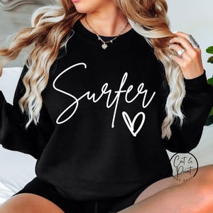 Puede incluir: Sudadera negra con la palabra "Surfer" en escritura blanca y un coraz&oacute;n blanco. La sudadera es de cuello redondo. La imagen tambi&eacute;n incluye el texto "Cat & Print SVG SHOP".