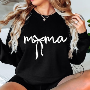 Puede incluir: Sudadera negra con la palabra "momma" en escritura blanca y un diseño de lazo. La sudadera es de cuello redondo. La imagen también incluye el logotipo "Cat & Print SVG Shop".