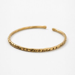 May include: A gold-coloured open cuff bracelet. The bracelet has a textured surface with a repeating pattern of small, leaf-like shapes. The bracelet is made of metal and is displayed on a white background.