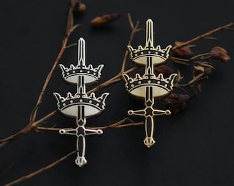 May include: Two enamel pins featuring a sword with three crowns on top. The pins are in different colours, one is silver and the other is gold. The pins are on a branch with dried leaves.