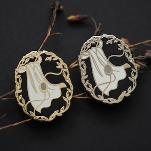 Peut inclure: Deux épingles ovales avec un fond noir et une bordure dorée. Chaque épingle présente un motif blanc avec un centre gris et une bordure de couronne de laurier dorée. Les épingles sont posées sur un fond sombre avec des brindilles sèches.