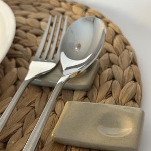 May include: A stainless steel fork and spoon rest on a small, rectangular, grey ceramic dish. The utensils are placed on a woven, light brown placemat. A white plate is partially visible in the background.