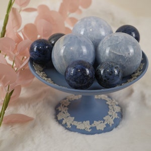 May include: A blue and white ceramic pedestal cake stand with a decorative floral design. The stand is filled with six smooth, round, blue and white crystal spheres.