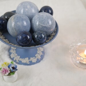 May include: A blue and white ceramic pedestal with a collection of blue and gray crystal spheres. The pedestal is decorated with a floral design. A small white ceramic flower pot with a bouquet of flowers sits in front of the pedestal. A clear glass candle holder with a lit candle sits on the right side of the image.