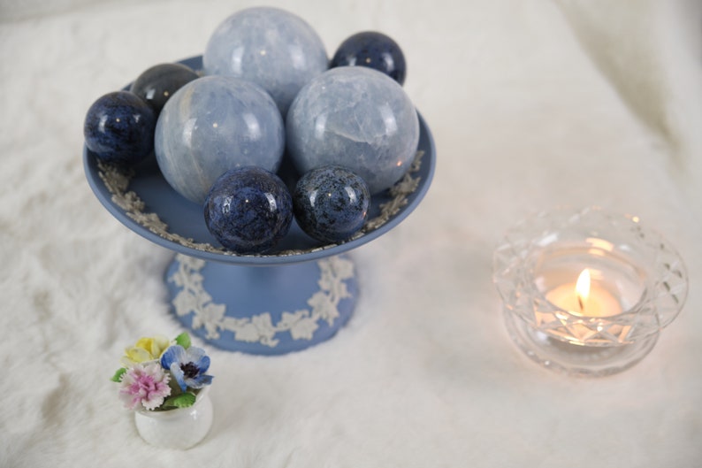May include: A blue and white ceramic pedestal with a collection of blue and gray crystal spheres. The pedestal is decorated with a floral design. A small white ceramic flower pot with a bouquet of flowers sits in front of the pedestal. A clear glass candle holder with a lit candle sits on the right side of the image.