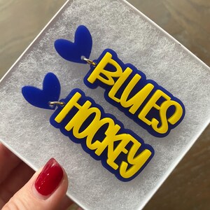 May include: A pair of blue and yellow earrings with the words "Blues Hockey" on them. The earrings are shaped like hearts and have a gold hook.