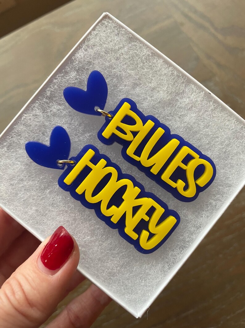 May include: A pair of blue and yellow earrings with the words "Blues Hockey" on them. The earrings are shaped like hearts and have a gold hook.