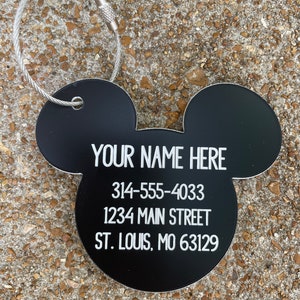 May include: Black Mickey Mouse-shaped tag with a silver metal ring and cable. The tag has white text that reads "YOUR NAME HERE", "314-555-4033", "1234 MAIN STREET", and "ST. LOUIS, MO 63129".