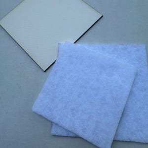May include: A light-colored, square wooden coaster with a dark brown edge sits next to two light blue, square felt pads. The items are on a light gray surface. The coaster and pads are likely for home decor or crafting.