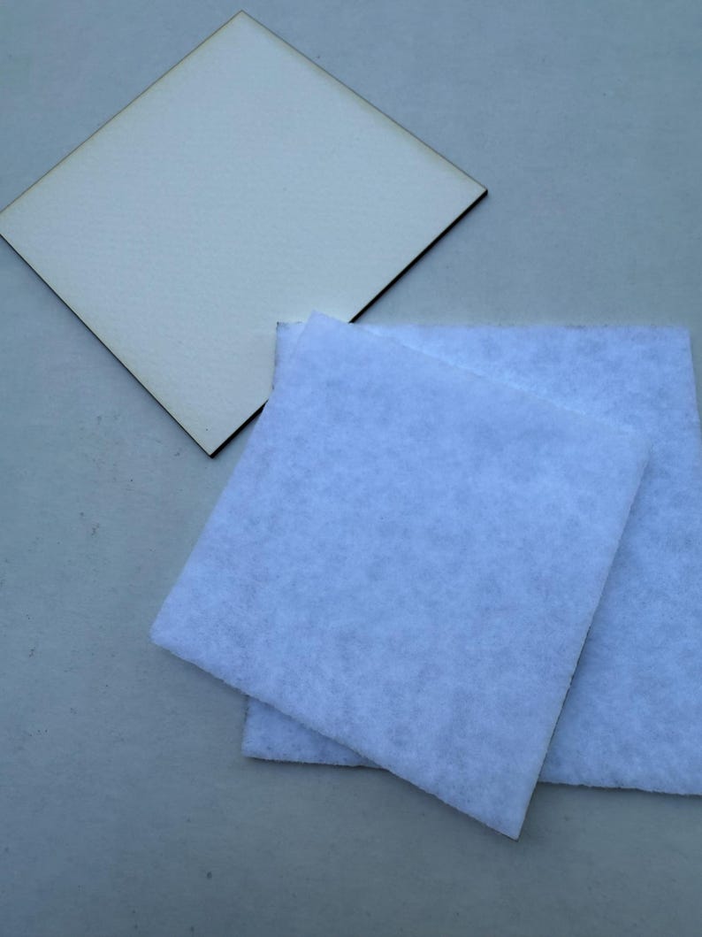 May include: A light-colored, square wooden coaster with a dark brown edge sits next to two light blue, square felt pads. The items are on a light gray surface. The coaster and pads are likely for home decor or crafting.