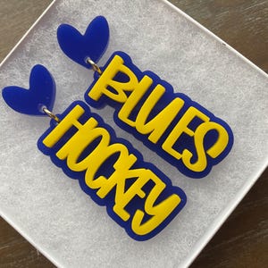 May include: A pair of blue and yellow acrylic earrings with a heart charm and the words "Blues Hockey" on each earring.