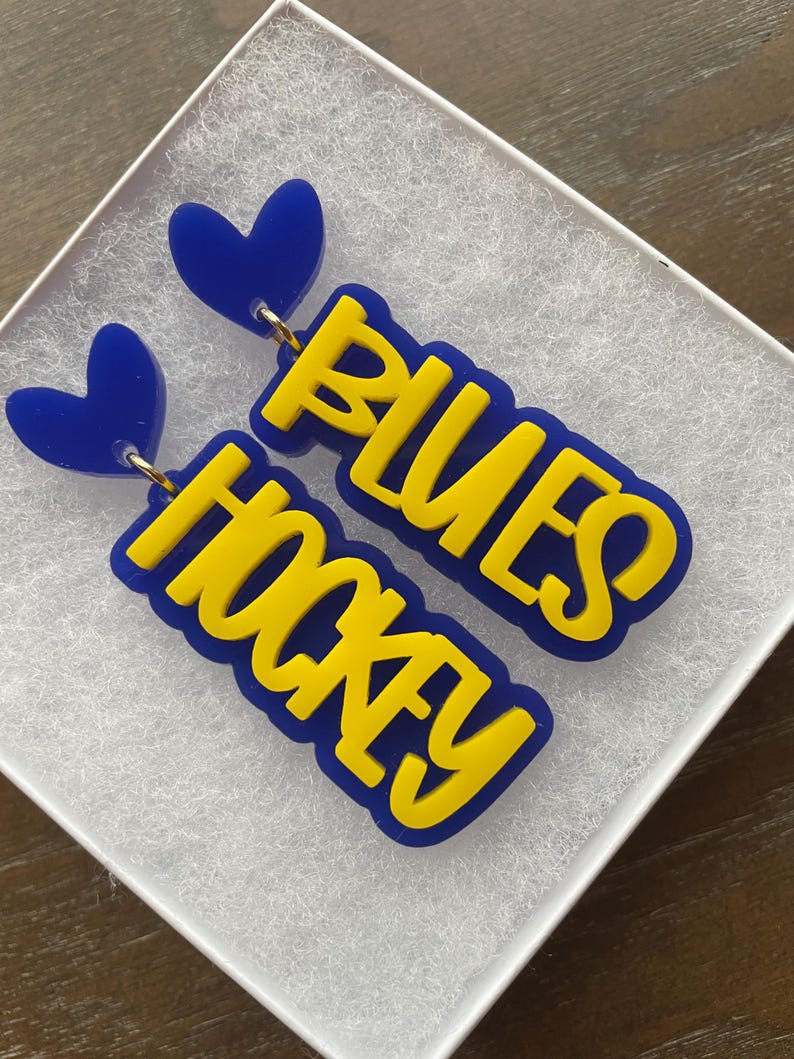 May include: A pair of blue and yellow acrylic earrings with a heart charm and the words "Blues Hockey" on each earring.