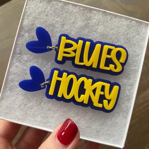 May include: A pair of blue and yellow acrylic earrings with the words "Blues Hockey" and a blue heart charm on each earring.