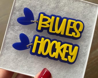 May include: A pair of blue and yellow acrylic earrings with the words "Blues Hockey" and a blue heart charm on each earring.