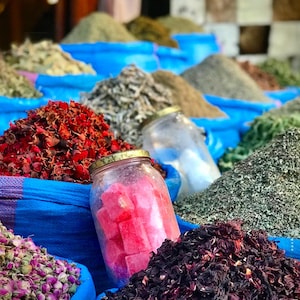 May include: A vibrant market display of various spices and herbs. Piles of dried red petals, green herbs, and dark purple spices are arranged in blue sacks. A glass jar filled with pink, cube-shaped items sits among the spices, suggesting a selection of culinary ingredients.