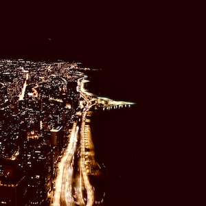 Puede incluir: Vista aérea nocturna de una ciudad iluminada. Las luces doradas de edificios y carreteras contrastan con el cielo oscuro. La costa es visible, con las luces de la ciudad reflejadas en el agua. La imagen captura la belleza de una ciudad de noche.