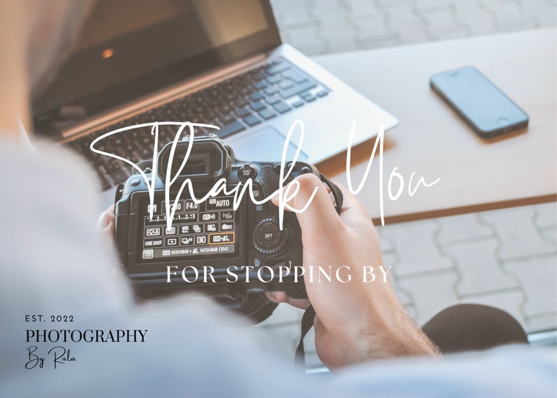 May include: A person holding a black DSLR camera with the text "Thank You for Stopping By" on the screen. The camera is in focus, and the person's hand is blurred. The background is out of focus and shows a laptop and a cell phone.