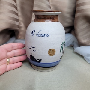 Könnte beinhalten: Eine weiße Keramikvase mit braunem Rand und einem blauen und grünen bemalten Design. Die Vase zeigt einen Wal, Vögel, eine Sonne und eine Palme. Der Text "Pto. Vallarta" ist auf der Vase geschrieben.