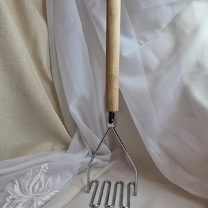 Puede incluir: Un pasapurés con mango de madera y cabezal de acero inoxidable. El mango es de color marrón claro con un agujero en la parte superior. El cabezal tiene un diseño ondulado. El utensilio de cocina está sobre un fondo blanco y beige.