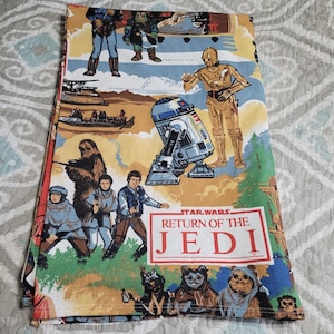 Peut inclure: Un tissu plié sur le thème de Star Wars Le Retour du Jedi. Le tissu présente des illustrations colorées de personnages tels que R2-D2, C-3PO, Chewbacca et les Ewoks. Le titre "Star Wars Le Retour du Jedi" est affiché dans un encadré rouge et blanc.