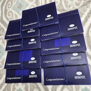 May include: A pile of navy blue photo albums, secured with blue elastic bands. Each album displays the text "Congratulations!" and the "memories" logo in white. The background is a patterned fabric in shades of grey and white.