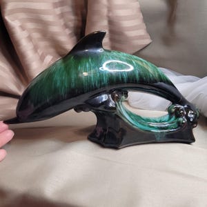 Puede incluir: Figura de delfín de cerámica en tonos verdes y negros. El delfín está arqueado, con su cola y aleta dorsal visibles. La figura está sobre una base que se asemeja a una ola. El delfín mide aproximadamente 30 cm de largo.