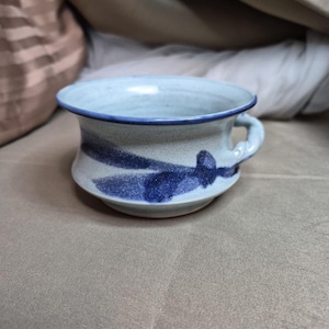 Peut inclure: Un bol en céramique avec une finition bleu clair mouchetée et un bord bleu foncé. Le bol est doté d'une petite anse et est décoré de coups de pinceau bleu foncé. Le bol est probablement fait à la main et convient pour servir des soupes ou d'autres plats.