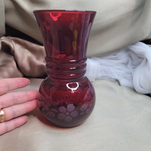 Peut inclure: Un vase en verre rouge foncé avec une large ouverture et une base ronde. Le vase présente un motif texturé autour du milieu et un motif floral gravé au fond.