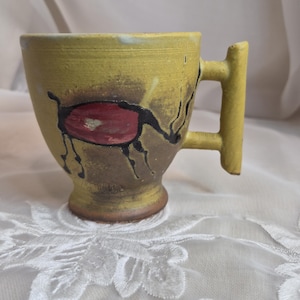 May include: A yellow ceramic mug with a textured surface and a pedestal base. The mug features a hand-painted black and red abstract animal figure on the side. It has a distinct, angular, flat-sided handle extending from the right side of the cup.