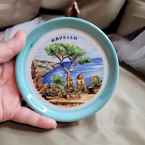Może przedstawiać: Mały, okrągły ceramiczny talerz z jasnoniebieską obwódką. Środek przedstawia malowniczą ilustrację Ravello we Włoszech, z drzewem, budynkami i morzem. Słowo "RAVELLO" jest wydrukowane u góry.