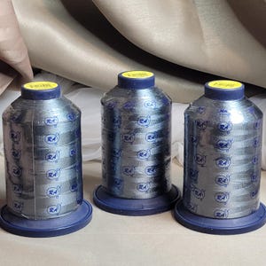 Puede incluir: Tres bobinas de hilo plateado metálico con bases y tapas azules. Cada bobina está envuelta en plástico transparente con un logotipo azul repetido. Las bobinas son cilíndricas y se asientan sobre una base plana y redonda.