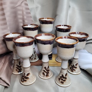 May include: A collection of eight ceramic goblets. Each goblet features a beige base, a dark brown rim, and floral patterns on the stem. The goblets are displayed against a neutral backdrop.