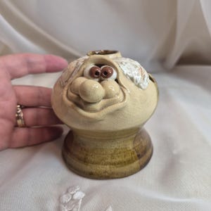 May include: A ceramic candle holder featuring a sculpted face with a large nose, wide smile, and two brown eyes. The upper portion is a light cream colour with a crackled glaze, while the base is a darker, mottled brown with a smooth finish.