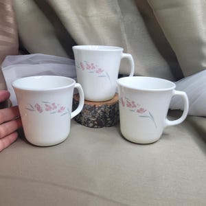 May include: Three white ceramic mugs with a floral design in pink and gray. Each mug has a handle and a curved shape. The mugs are arranged on a neutral surface.
