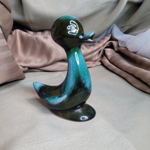 Puede incluir: Una figurita de pato de cerámica esmaltada en verde y azul. El pato tiene un cuello largo y un cuerpo redondeado. El esmalte es verde y azul moteado, con un ligero brillo.