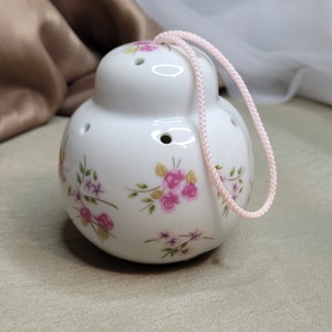 以下が含まれることがあります： ピンクの花柄が描かれた白い陶器製のポット。ピンクの紐の取っ手が付いています。ポットには通気用の小さな穴が開いています。