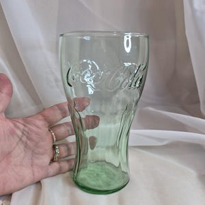 May include: A light green, translucent glass tumbler featuring the embossed Coca-Cola logo in script lettering. The glass has a classic contoured shape with vertical fluted ridges along the lower section and a smooth, wide rim at the top.