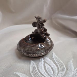 May include: A small, brown ceramic ashtray with a decorative dragon figurine on top. The ashtray has a circular shape with a detailed rim. The dragon is dark brown and has a whimsical design. The ashtray is placed on a white surface.