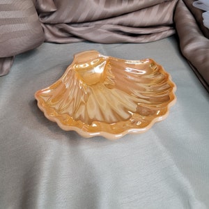 Può includere: Un piatto iridescente a forma di conchiglia con un bordo smerlato. Il piatto ha una superficie lucida e riflettente con sfumature di arancione e oro. Il pezzo decorativo è probabilmente in vetro o ceramica.