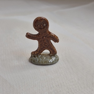 Peut inclure: Petite figurine en céramique représentant un bonhomme en pain d'épices brun, souriant, avec trois boutons. La figurine est posée sur un petit socle vert. Objet décoratif fantaisiste.