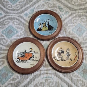 May include: Three decorative plates with wooden frames. Each plate features a different vintage illustration in a circular design. The illustrations depict scenes with people and text. The plates are arranged in a triangular pattern.
