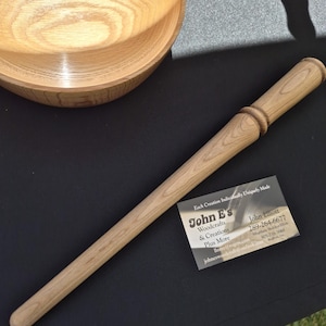 May include: A light-coloured wooden wand with a rounded tip and a decorative ring near the handle. A wooden bowl is in the background. A business card with the text "John E's Woodcrafts & Creations" is also visible.
