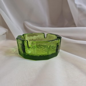 Pode incluir: Um cinzeiro de vidro verde-limão com um exterior espesso e facetado e um interior liso. O recipiente circular apresenta quatro entalhes distintos ao longo da borda, concebidos para apoiar objetos, e exibe uma cor verde vibrante e translúcida.