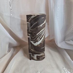 May include: A cylindrical ceramic vase featuring a marbled pattern of dark brown and white swirls. The exterior surface has a textured, matte finish with three distinct vertical rectangular indentations along one side of the vessel.
