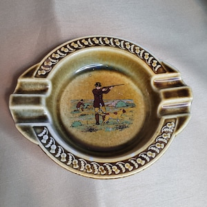 Puede incluir: Un cenicero de cerámica verde y marrón con un diseño de cardos en relieve alrededor del borde. El centro presenta una escena de un hombre cazando con un perro.