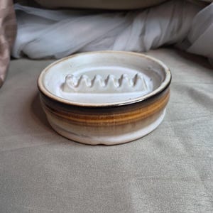 May include: A ceramic soap dish with a circular design. The dish features a white interior with a ridged pattern and a brown and tan exterior. The dish is designed to hold a bar of soap.